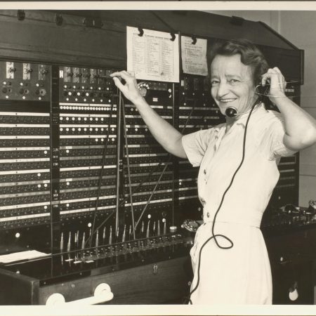Black at white photograph of female standing at telephone switchboard, smiling at camera.
