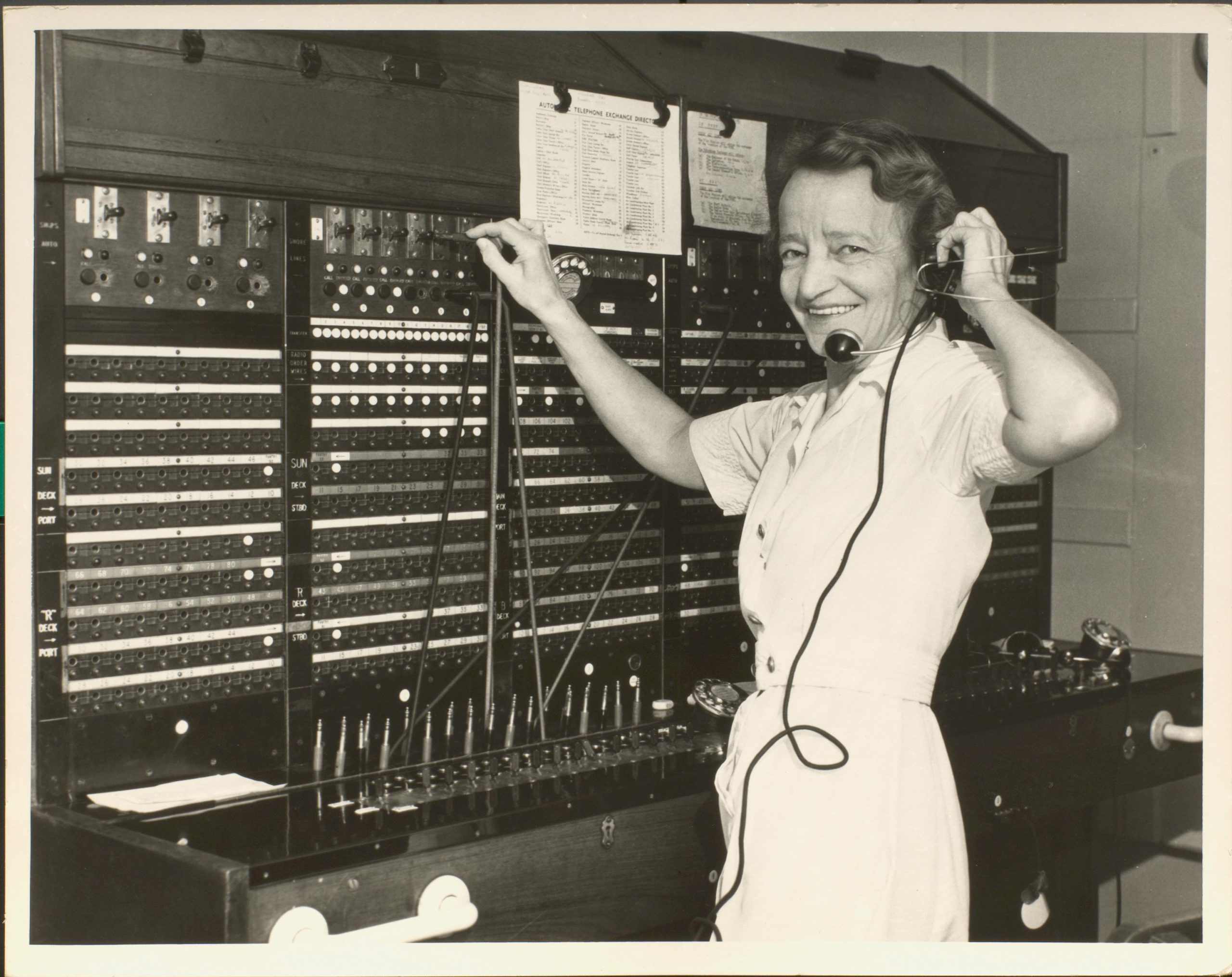Black at white photograph of female standing at telephone switchboard, smiling at camera.