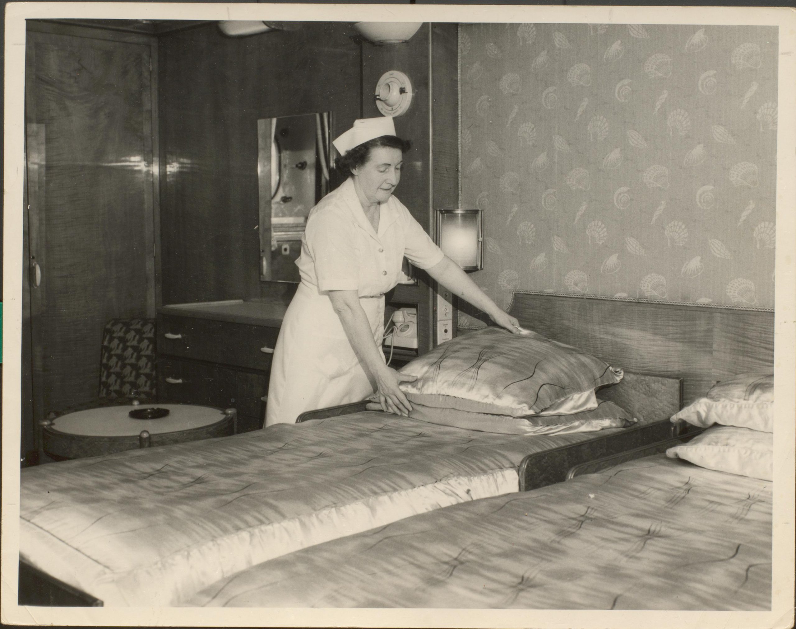 Black and white photograph of female in uniform arranging pillows on a single bed in a cabin.