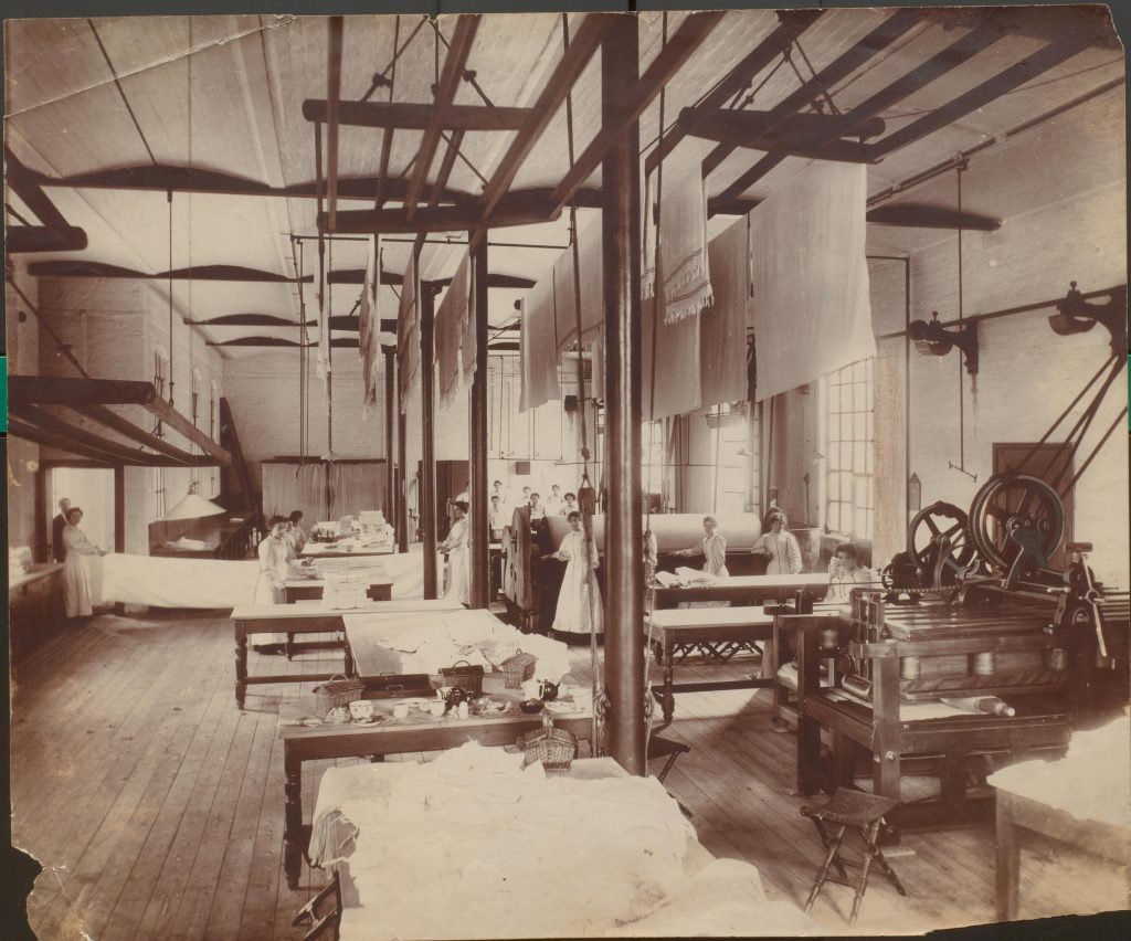 Sepia photograph showing women working in the laundry and mangling department. The scene includes fabric hanging from the ceiling to dry, women sat at a large mangle, and women folding large pieces of cloth.
